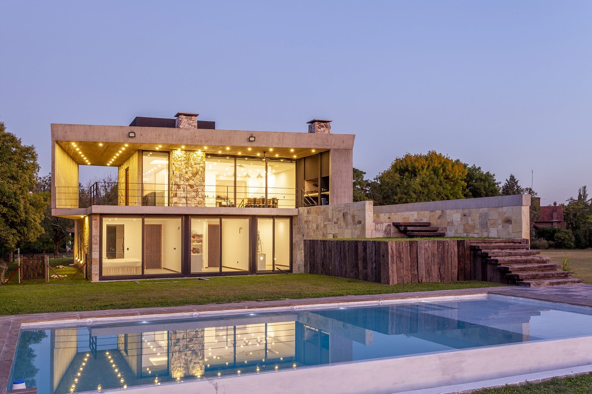Casa de piedra y hormigón iluminada al anochecer, con la piscina en primer plano